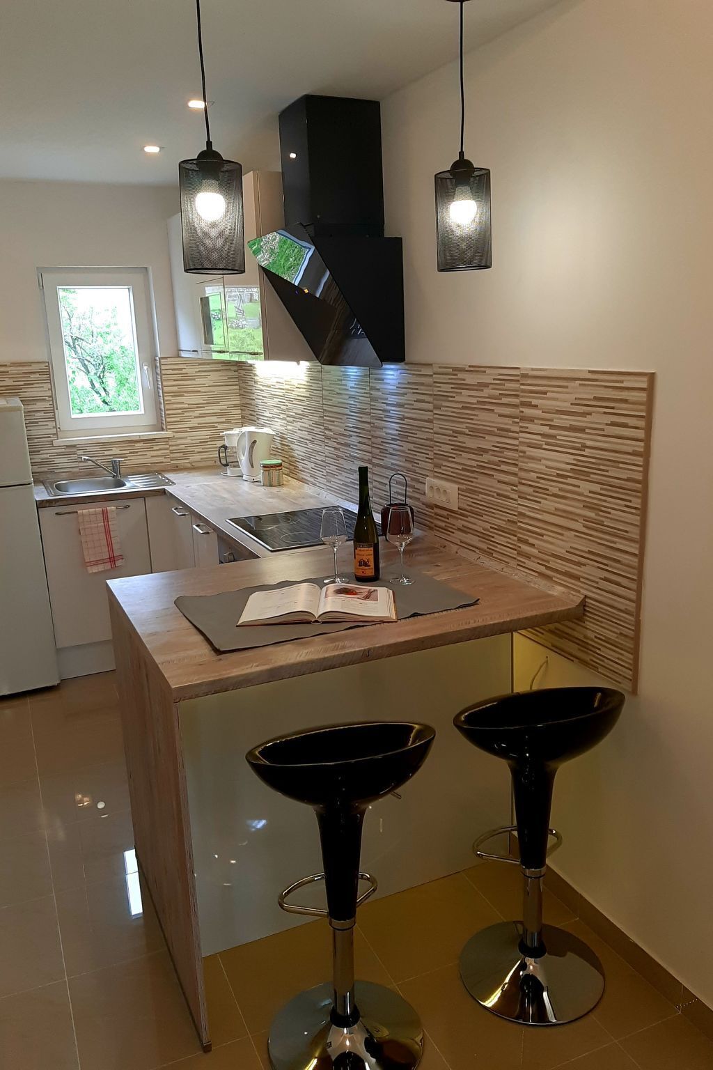 A modern kitchen with a central island, two bar stools, hanging pendant lights, and a window with a view of greenery.