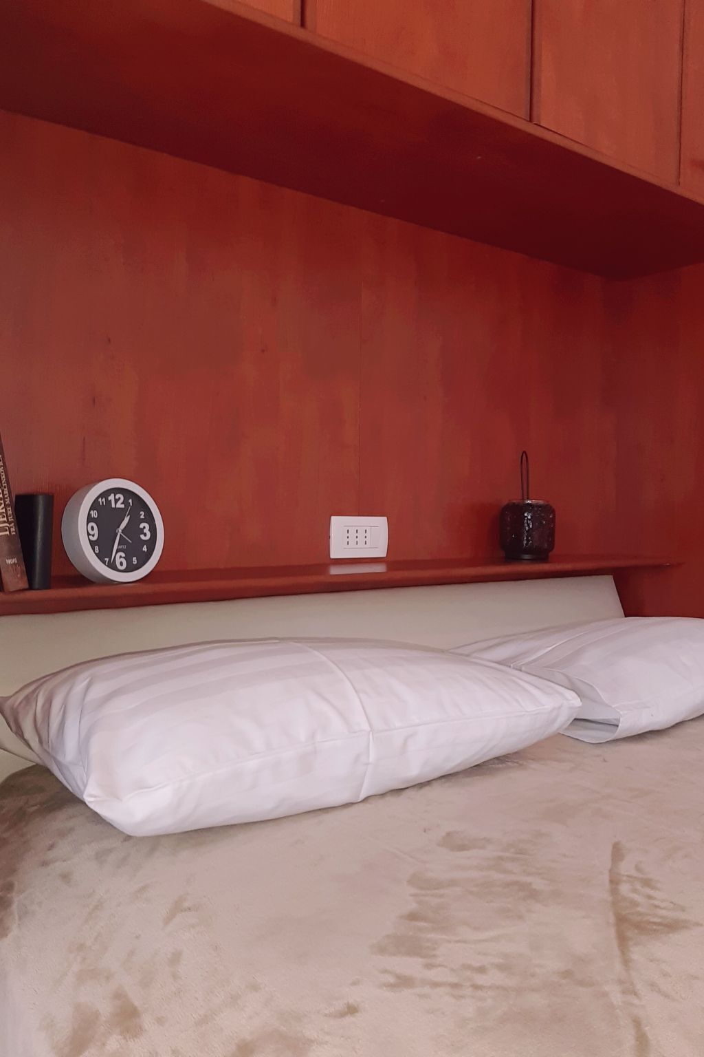A bed with white bedding, a clock, a power outlet, and a small black object on a wooden shelf.