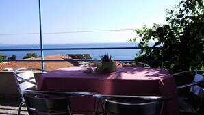 Ein Tisch mit kariertem Tischläufer, zwei Stühle und ein Blumentopf auf einem Balkon mit Blick aufs Meer.