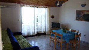 Ein Zimmer mit Holzboden, einer Couch, einem Esstisch mit Stühlen, einem Fernseher und einem Fenster mit Vorhängen.