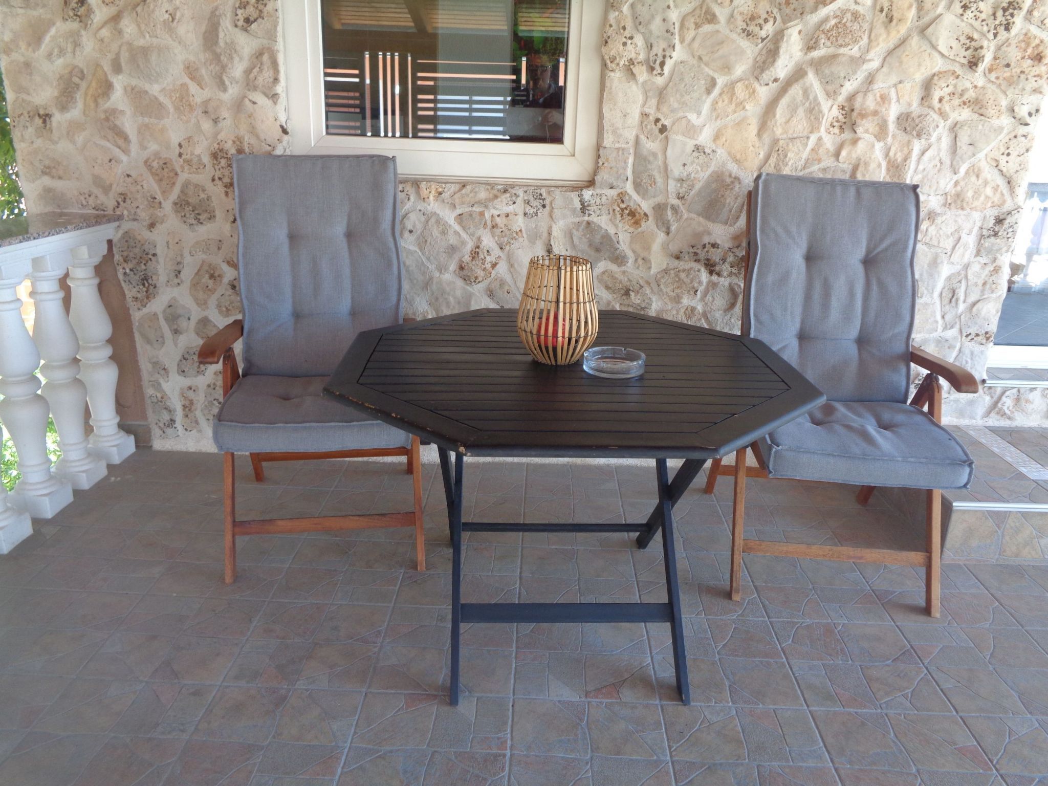 A stone wall, a window, two grey upholstered chairs, a round table, and a woven basket with a glass dish.