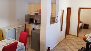 Eine kleine Küche mit Gaskochfeld, Kaffeemaschine und Kühlschrank. Es gibt einen Esstisch mit Stühlen und einen kleinen Tisch mit einem farbenfrohen Gegenstand darauf.