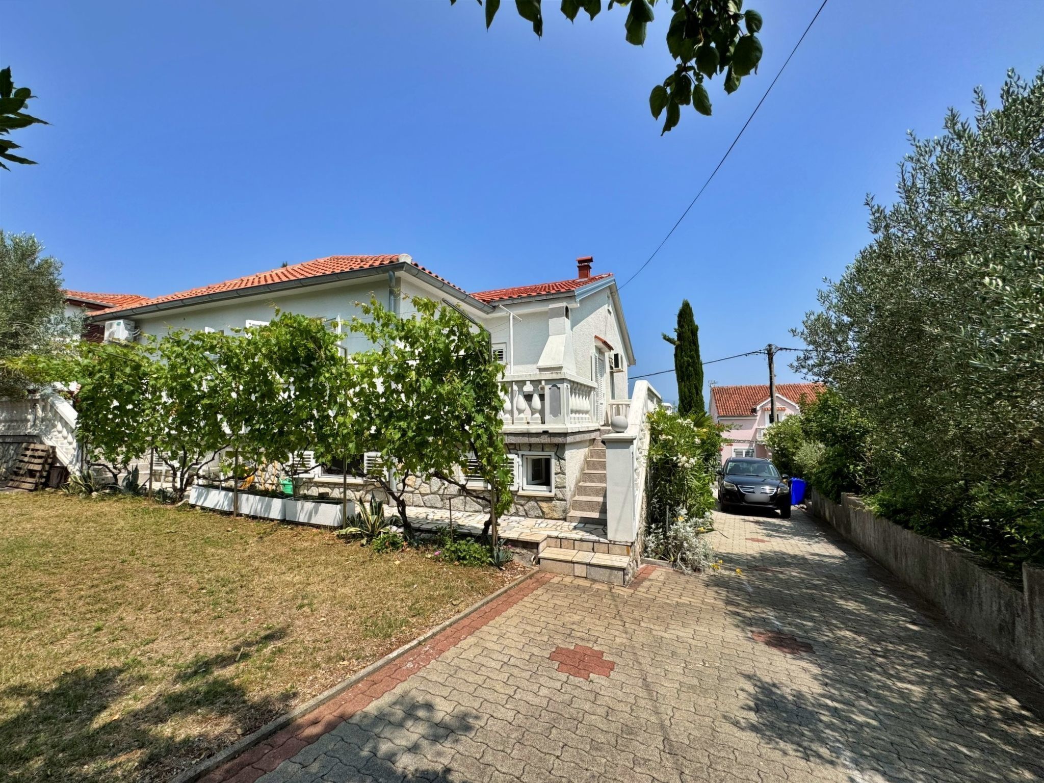 A two-story house with a red-tiled roof, a front garden with trees, and a paved driveway.