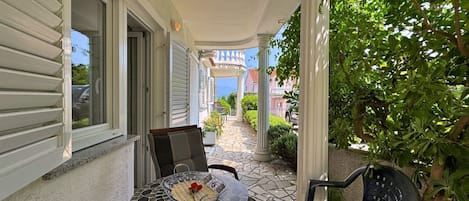 Um alpendre coberto com mesa e cadeiras azulejadas, vista para a rua e uma planta verde exuberante.
