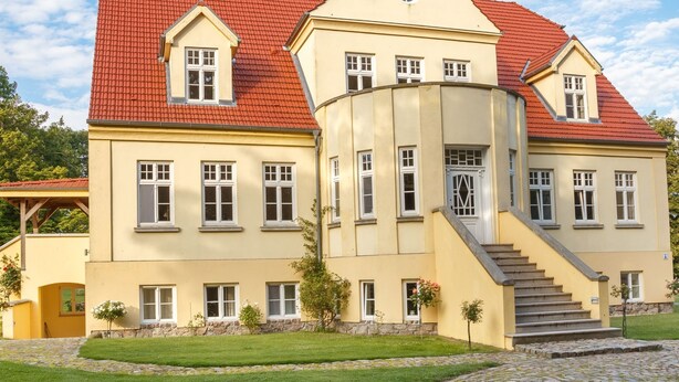 Ein großes, gelbes Gebäude mit rotem Dach, zahlreichen Fenstern und einer Treppe, die zum Eingang führt.