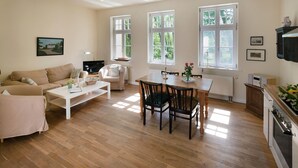 Ein geräumiges Wohnzimmer mit einer Couch, einem Couchtisch und einem Essbereich. Große Fenster sorgen für natürliches Licht und Blick ins Grüne.