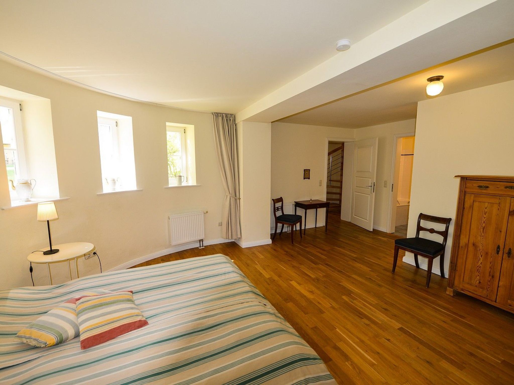 A bedroom with a bed, a desk, and a chair. There is a radiator, a window, and a door leading to another room.