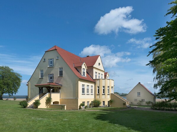 Une grande maison à deux étages, avec un toit recouvert de tuiles rouges, un balcon et une pelouse bien entretenue.