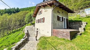 Ein kleines, rustikales Chalet mit einer Steintreppe, die zu einer Holzterrasse führt, umgeben von üppiger Vegetation und einem bewaldeten Hügelhang.