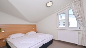 Un dormitorio con cama, cabecera, ventana con cortinas, radiador e iluminación fijada a la pared.