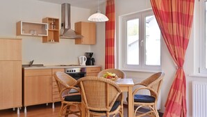 Cocina compacta con armarios de madera, estufa a gas y cafetera. Cuenta con una zona de comedor con sillas de mimbre y una mesa, además de una ventana con cortinas.