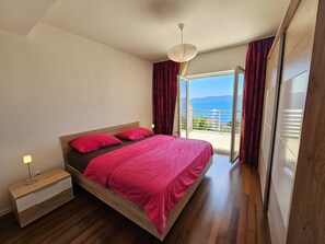 A bedroom with a wooden bed, red bedding, a nightstand, and a view of the sea through a glass door. - Wohnung für 6 Personen mit Meerblick by Interhome (Novi Vinodolski)