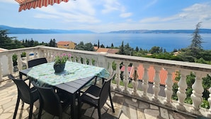 Eine Terrasse mit Tisch und Stühlen, ein gestreifter Sonnenschirm und Blick auf einen See und Berge.