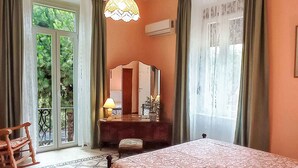 Een slaapkamer met een bed, een dressoir, een spiegel, een lamp en uitzicht op groen buiten het raam.
