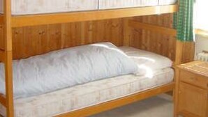 A bunk bed with two levels, wooden walls, a small bedside table, and a window with green curtains. - Suot Rövens by Interhome (Samedan)