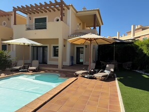 Zona de piscina con sillones de descanso y sombrillas, una pérgola y un edificio con techo de tejas.