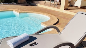 En solstol vid poolkanten med en handduk och solglasögon, ett bubbelbad och en uteplats med kakel och möbler i rotting.