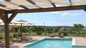 Un jacuzzi bajo una pérgola, con una piscina y sillones de descanso al fondo.