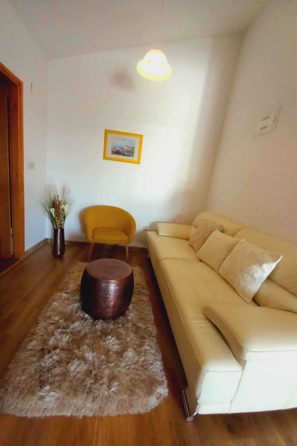A living room with a beige sofa, a round wooden coffee table, a yellow armchair, and a framed picture on the wall.