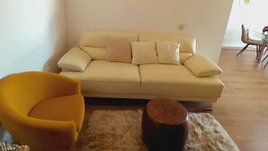 Un salón con un sofá, un sillón amarillo, una mesa de centro redonda de madera y una alfombra mullida.