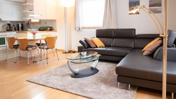 A modern living room with a glass coffee table, a black leather sofa, and a dining area with a white kitchen in the background. - Chesa La Capriola by Interhome (Sils Maria)