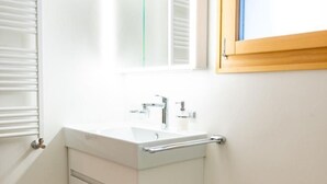 A modern bathroom with a white vanity, a mirror, a window with blinds, and a trash bin. - Chesa La Capriola by Interhome (Sils Maria)