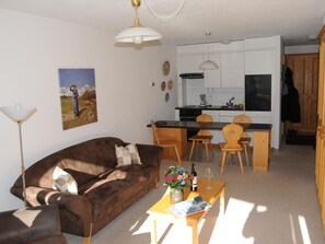 Ein gemütliches Wohnzimmer mit einem braunen Sofa, einem Esstisch mit Stühlen, einer Küchenzeile mit weißen Schränken und einer Pendelleuchte.