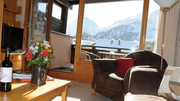 Un accogliente soggiorno con un tavolo in legno, una poltrona e una sedia. Un'ampia finestra offre vista su montagne innevate.
