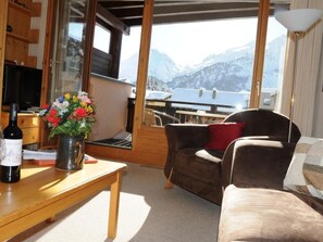 Ein gemütliches Wohnzimmer mit einem Holztisch, einer Couch und einem Sessel. Ein großes Fenster bietet Blick auf schneebedeckte Berge.