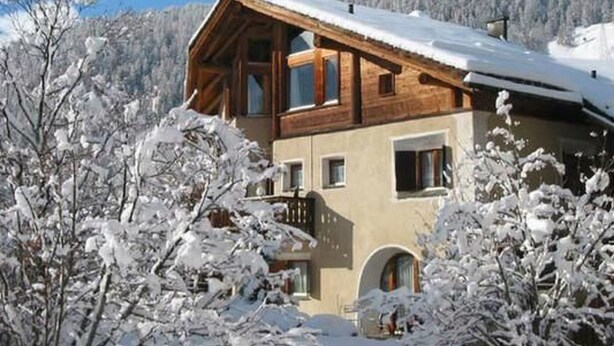 Una baita di legno con un balcone, circondata da alberi e montagne innevate.