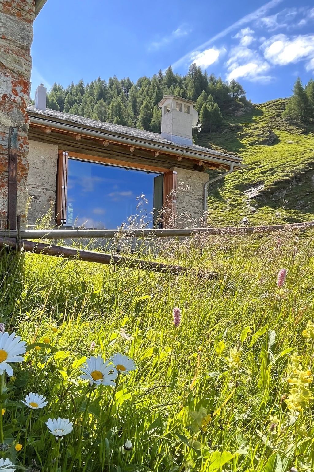 Un bâtiment en pierre doté d’une grande fenêtre et d’une terrasse en bois, entouré d’un champ verdoyant parsemé de fleurs sauvages.