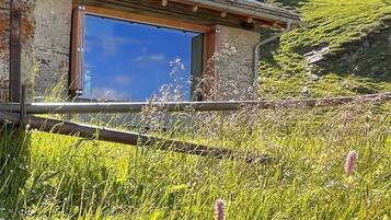 Un bâtiment en pierre doté d’une grande fenêtre et d’une terrasse en bois, entouré d’un champ verdoyant parsemé de fleurs sauvages.