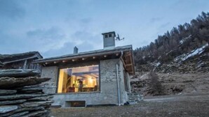 Eine Steinhütte mit großem Fenster, umgeben von einer Steinmauer und einem bewaldeten Hang.