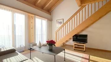Ein geräumiges Wohnzimmer mit hoher Holzbalkendecke, einer Treppe, einem Fernseher auf einem Schrank und einem großen Fenster mit transparenten Vorhängen.