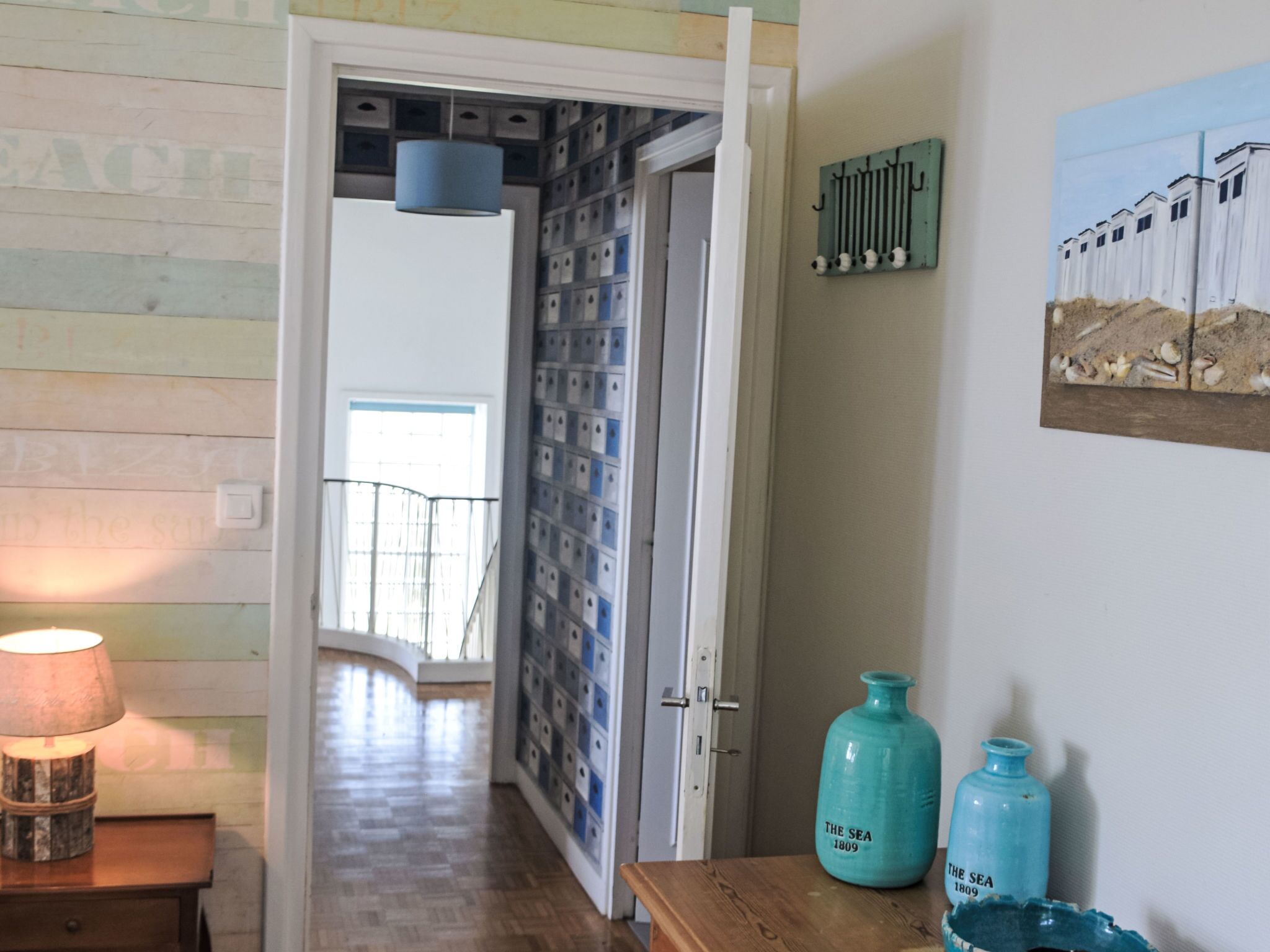 A hallway with a wooden floor, a door leading to a room with a blue lamp, and two blue containers on a table.
