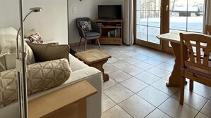 Ein Wohnzimmer mit einer Couch, einem Fernseher, einem Sessel, einem Couchtisch und einem großen Fenster mit Blick auf Bäume.