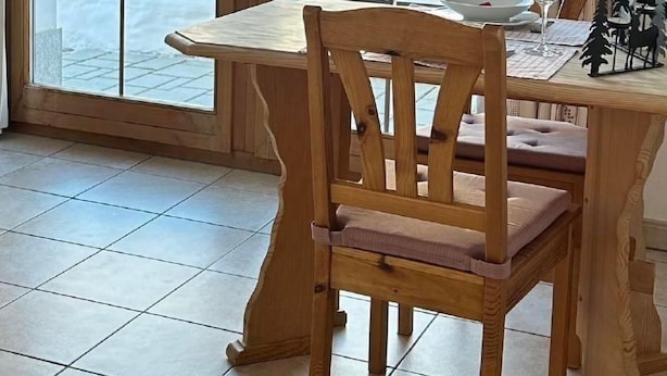 Ein Essbereich mit einem Holztisch und Stühlen, einem Fenster mit Blick auf Bäume und eine verschneite Landschaft sowie einer Lampe auf dem Tisch.