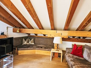 Un salon de style industriel, avec des poutres en bois apparentes, un canapé d’angle, un téléviseur et une petite table avec une lampe.