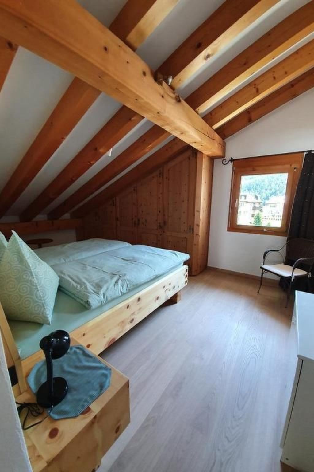 A small attic bedroom with a wooden ceiling, a single bed, a desk, and a window with a view of the outside.