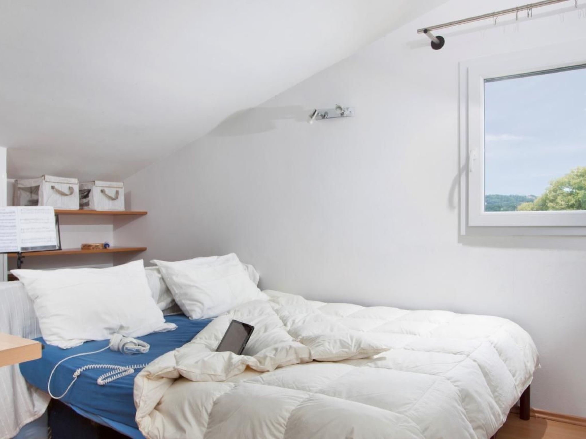 A bedroom with a bed, a bedside table, a shelf with storage boxes, a window with a view, and a thermostat on the wall.