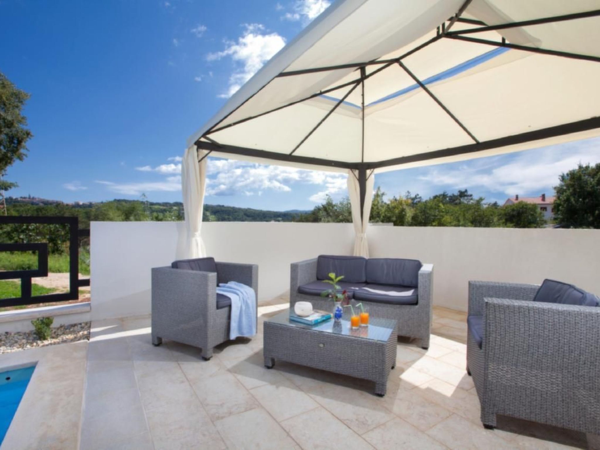 Un espacio de estar al aire libre cubierto con mobiliario de mimbre y una mesa de centro de vidrio, con vistas a una piscina y vegetación.