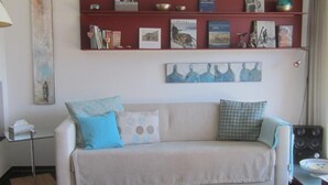 Sala de estar com sofá, almofadas, uma estante repleta de livros e objetos decorativos, e um quadro na parede.