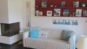 Uma sala de estar com um sofá branco, almofadas azuis e brancas, uma estante vermelha com livros e objetos decorativos, um quadro e um abajur.