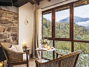 A cozy room with wicker furniture, a table with candles, and a view of mountains through large windows. - Ländliche Wohnung El Carbayu by Interhome (Las Ventas)