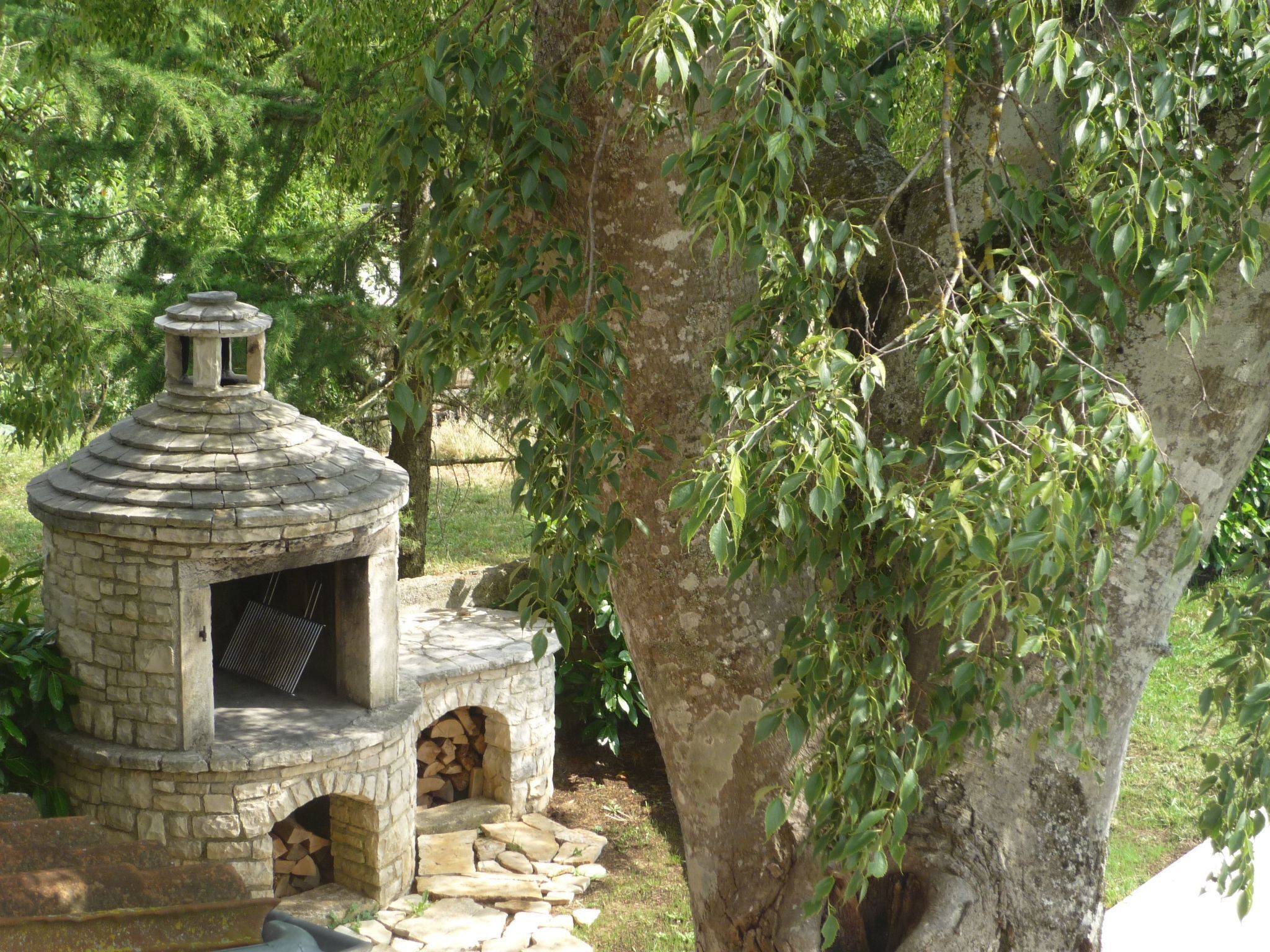Un four extérieur en pierre, doté d’une cheminée, entouré d’arbres et d’herbe.