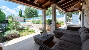 Ein überdachter Patio-Bereich mit Pool, Gartenmöbeln, Steinmauern und viel Grün.