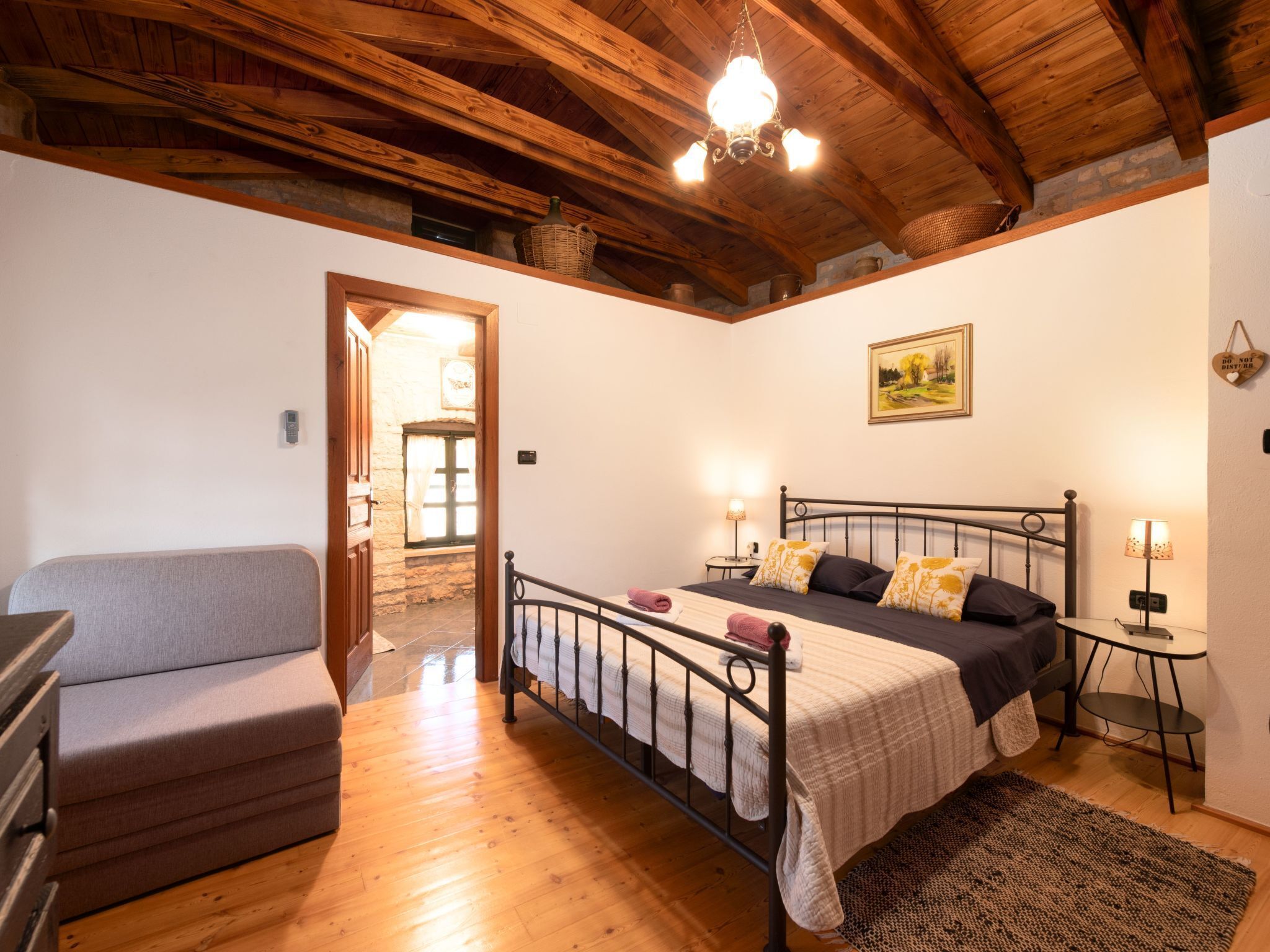 A bedroom with a wooden ceiling, a bed with a metal frame, a gray sofa, and a small table with a lamp.