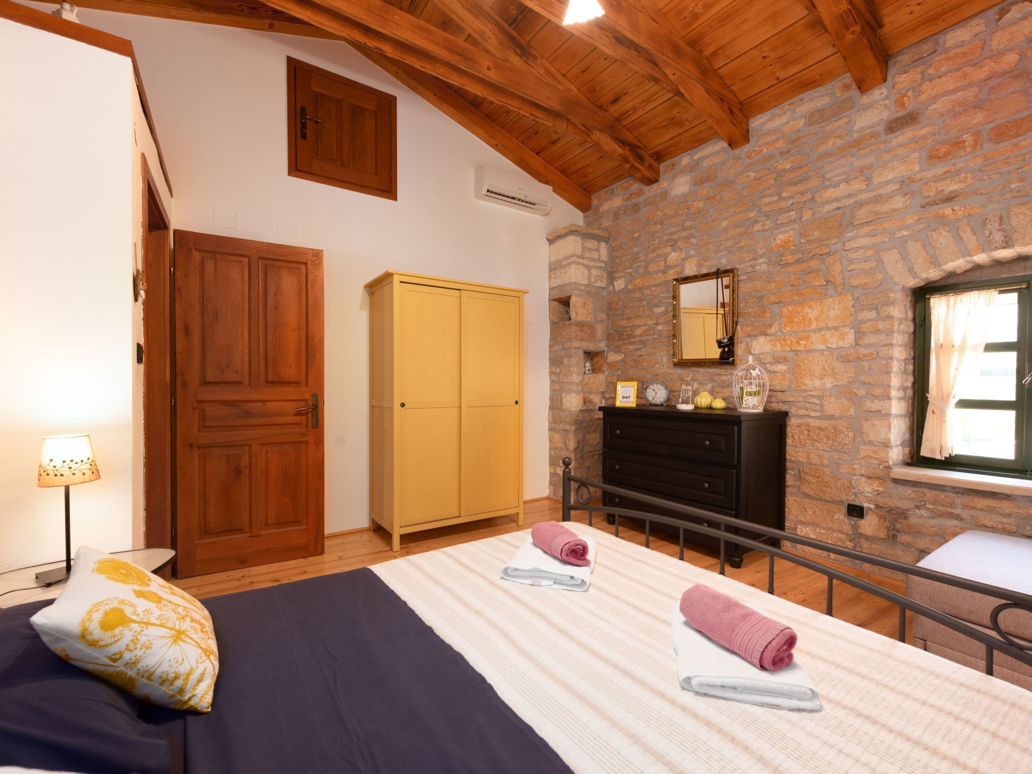Een slaapkamer met een houten plafond, een stenen muur, een bed met een gestreept dekbedovertrek, een houten garderobe, een dressoir met spiegel, en een raam met gordijnen.