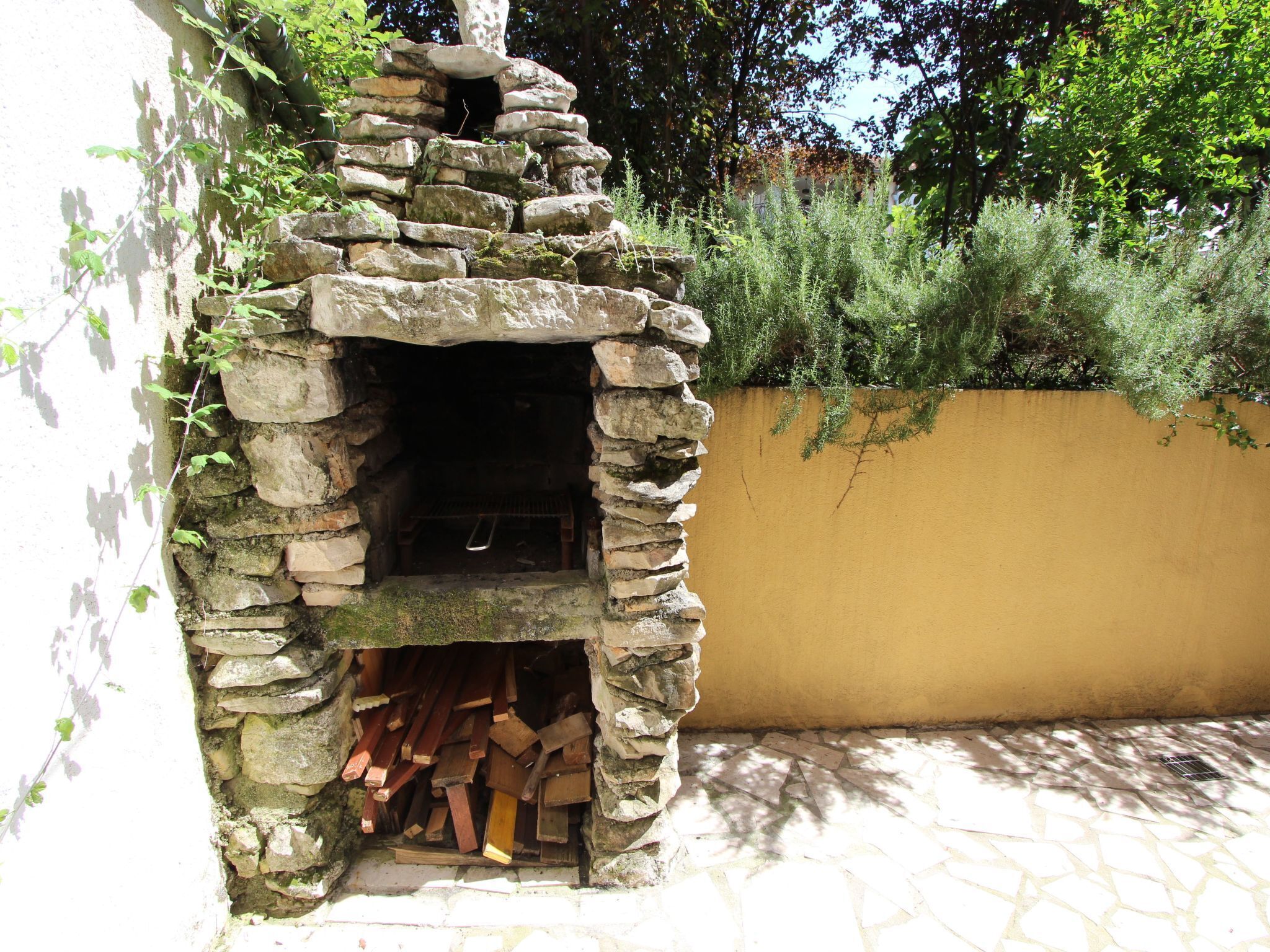 A stone-built outdoor fireplace with logs stored inside, surrounded by greenery and a yellow wall.