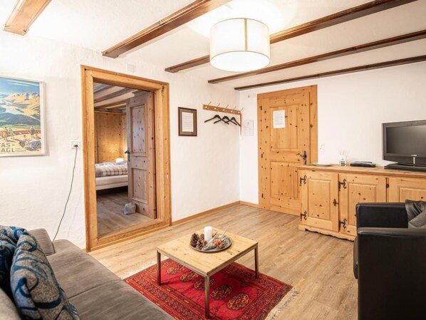 A cozy living room with a sofa, a coffee table, and a TV. There's a wooden door leading to a bedroom, and a red rug on the floor.